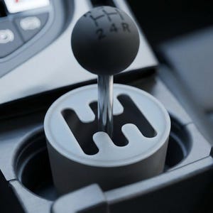 May include: A close-up of a car's gear shift. The shift knob is black and round, with the gear positions 1, 2, 3, 4, and R printed in white. The shift is set in a white and gray housing, with a silver metal shaft.