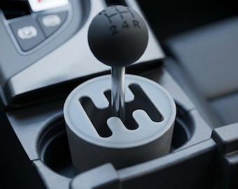 May include: A close-up of a car's gear shift. The shift knob is black and round, with the gear positions 1, 2, 3, 4, and R printed in white. The shift is set in a white and gray housing, with a silver metal shaft.