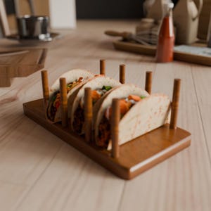 May include: A wooden taco holder with four tacos. The tacos are filled with various ingredients, including what appears to be meat, vegetables, and sauce. The holder is made of a dark wood and has vertical wooden supports to hold the tacos upright.