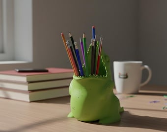 May include: A light green alligator-shaped pencil holder filled with various colored pencils. The holder sits on a wooden desk next to a stack of books, a mug, and a smartphone.