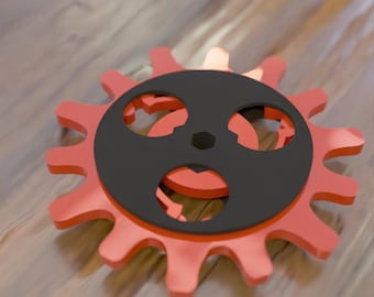 May include: A red and black gear with a circular design. The gear has a black center with three circular cutouts and is layered over a red gear with pointed teeth. The background is a light brown wooden surface.