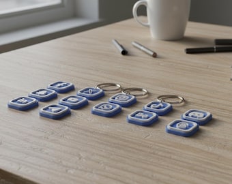 May include: A collection of blue and white keychains featuring social media icons, arranged on a light wood-grain surface. The keychains are square-shaped with silver key rings. A white mug, pens, and a book are also on the table.