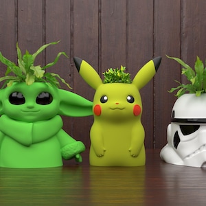 Puede incluir: Tres macetas temáticas de personajes: un Baby Yoda verde, un Pikachu amarillo y un casco de Stormtrooper blanco. Cada maceta contiene plantas verdes. Las macetas de Baby Yoda y Pikachu son verdes y amarillas. La maceta de Stormtrooper es blanca y negra.