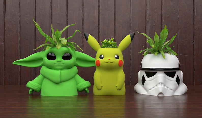 May include: Three character-themed planters: a green Baby Yoda, a yellow Pikachu, and a white Stormtrooper helmet. Each planter holds green plants. The Baby Yoda and Pikachu planters are green and yellow, respectively. The Stormtrooper planter is white and black.