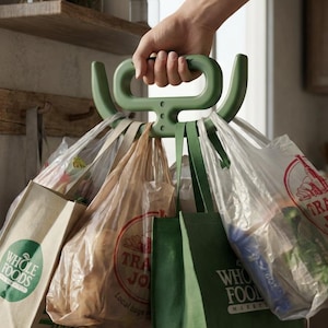 May include: A green plastic grocery bag carrier holds multiple shopping bags from Whole Foods and Trader Joe's. The bags are filled with groceries and are being carried by a person. The carrier has a handle for easy transport.