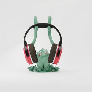 Puede incluir: Un soporte para auriculares verde con el tema de Bulbasaur que sostiene un par de auriculares negros y rojos. El soporte presenta una figura de Bulbasaur con brazos para sostener los auriculares. Los auriculares tienen orejeras marrones.