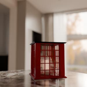 May include: A red wooden lantern shaped like a British phone booth. The lantern has a black roof and a glass enclosure with a candle inside. The lantern is sitting on a marble surface.
