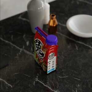 May include: A red and white carton with a purple cap. The carton features Japanese text and graphics. It sits on a dark marble surface, with white vases and a small gold bottle in the background.
