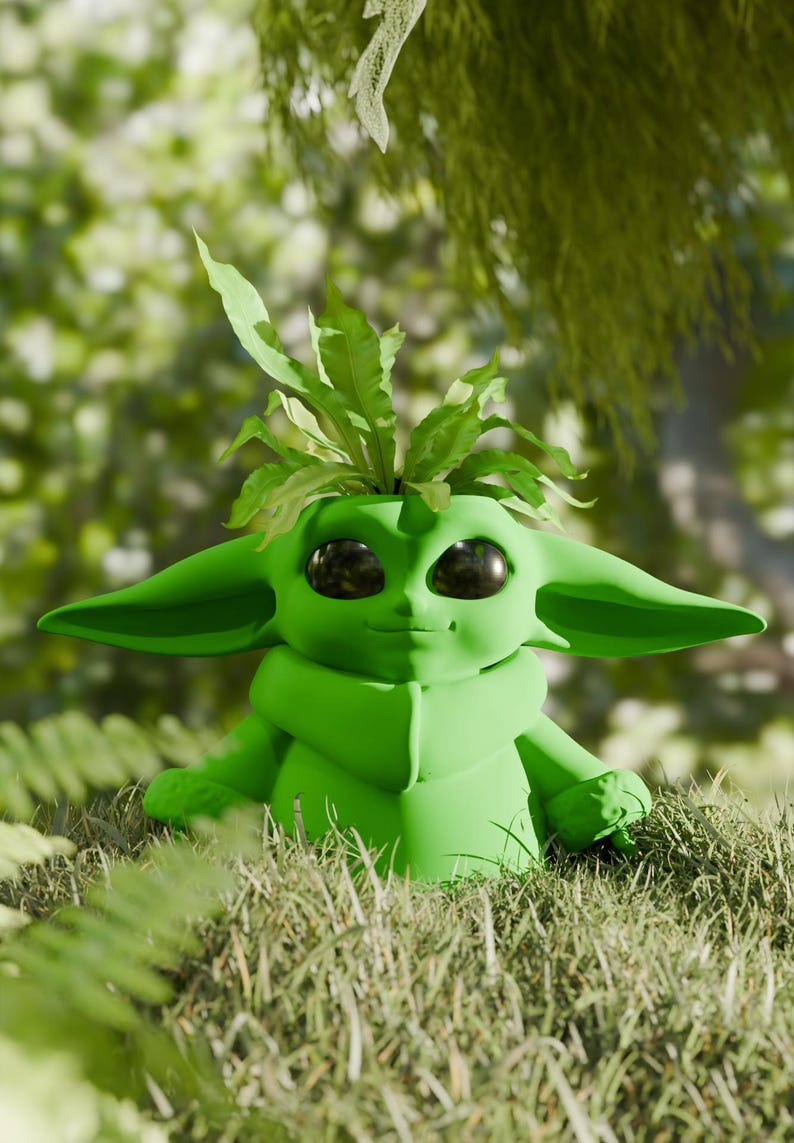 May include: A green Grogu-shaped planter with large ears and big eyes, filled with green leafy plants. The planter is sitting in a bed of grass, with a blurred green background of foliage.