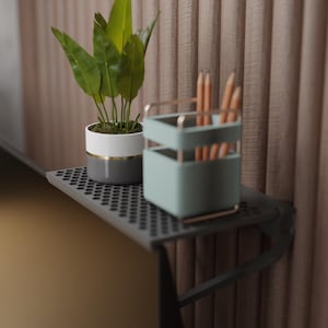 May include: A small, gray metal shelf with a perforated surface holds a potted plant and a desk organizer. The plant has large green leaves and is in a white, gray, and gold pot. The desk organizer is light blue and holds pencils.