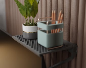 May include: A small, gray metal shelf with a perforated surface holds a potted plant and a desk organizer. The plant has large green leaves and is in a white, gray, and gold pot. The desk organizer is light blue and holds pencils.