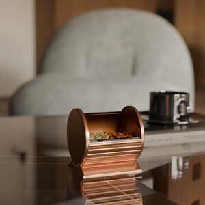 May include: A copper-colored, cylindrical container with horizontal bars and a rectangular base. The container holds small, colorful items. A black cup and saucer are in the background, resting on a book. The setting is a modern interior.