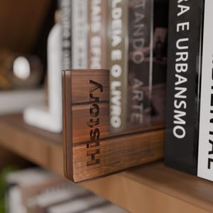 May include: A wooden bookend with the word "History" engraved vertically. The bookend is a warm brown color and sits on a wooden shelf next to a row of books. The bookend is designed to hold books upright.