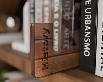 May include: A wooden bookend with the word "History" engraved vertically. The bookend is a warm brown color and sits on a wooden shelf next to a row of books. The bookend is designed to hold books upright.