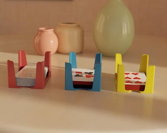 May include: Three colorful card holders in red, blue, and yellow, each holding a deck of playing cards. The holders are made of a solid material and have a simple, rectangular design with vertical supports. Decorative vases are in the background.