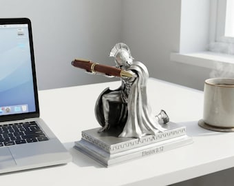 May include: A silver pen holder shaped like a kneeling Roman soldier holding a brown pen. The pen holder sits on a white desk next to a laptop and a mug of coffee. The base of the pen holder has the text "Ephesians 6:12".