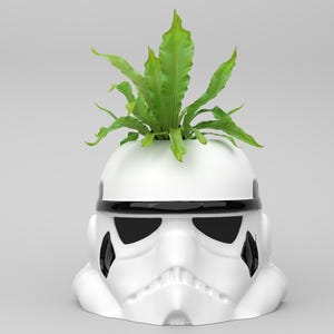 May include: A white ceramic planter shaped like a Stormtrooper helmet, with black accents and a green plant sprouting from the top. The planter is a unique decorative item for home or office.