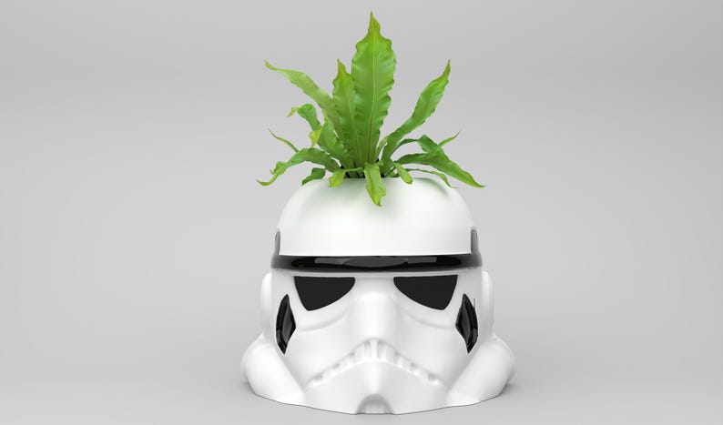 May include: A white ceramic planter shaped like a Stormtrooper helmet, with black accents and a green plant sprouting from the top. The planter is a unique decorative item for home or office.