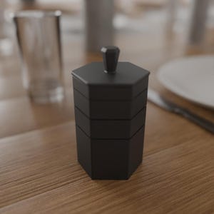 May include: A black, hexagonal, tiered container with a small knob on top. The container is made of a matte material and sits on a wooden table. A glass and a plate are in the background.
