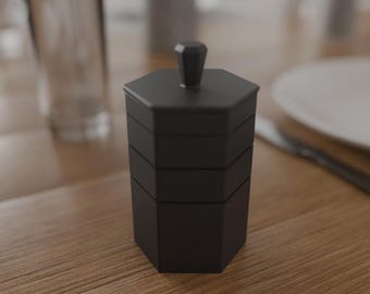 May include: A black, hexagonal, tiered container with a small knob on top. The container is made of a matte material and sits on a wooden table. A glass and a plate are in the background.