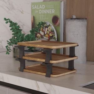 Puede incluir: Una bandeja de servicio de madera de tres niveles con soportes de color gris oscuro. Las bandejas son rectangulares con un acabado marrón claro. En el fondo se ve un libro de cocina titulado "SALAD for DINNER".