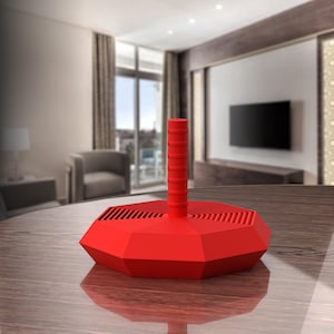 May include: A bright red, geometric-shaped object with a cylindrical handle. The base is a multi-sided polygon with a series of concentric lines. The handle is a series of stacked cylinders. The object is sitting on a wooden table.