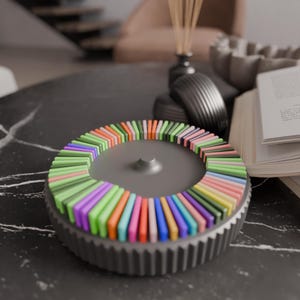 May include: A gray, gear-shaped decorative object with a circular arrangement of colorful, rectangular pieces. The pieces are in shades of green, purple, orange, and pink. The object sits on a dark, marble-like surface.
