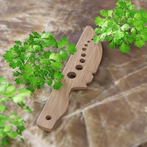 Könnte beinhalten: Ein Kräutermesser aus Holz mit einer Reihe kreisförmiger Löcher und einem Griff. Frische grüne Kräuter umgeben das Messer, was auf seine Verwendung zum Hacken von Kräutern hindeutet. Das Holz hat eine natürliche Maserung und eine hellbraune Farbe.