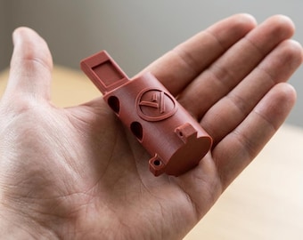 May include: A matte terracotta-colored 3D printed cylindrical object featuring a circular logo with a stylized letter V inside. The component has a rectangular tab extending from one end, two circular side openings, and small mounting flanges.