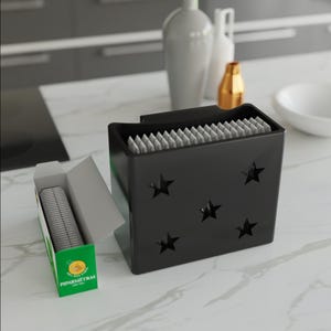 May include: A black, star-cutout container filled with individually wrapped items, next to a green and white box with the text "PIPARMETRAS". The container is on a white countertop, with decorative vases in the background.