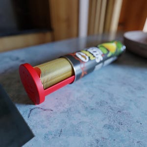 以下が含まれることがあります： 赤いベースと赤いスライド機構を備えた、ポテトチップスの円筒形容器。チップスは金色で、容器の中に積み重ねられています。容器には、ブランド名とライムと唐辛子のグラフィックが描かれた黒とシルバーのラベルが付いています。