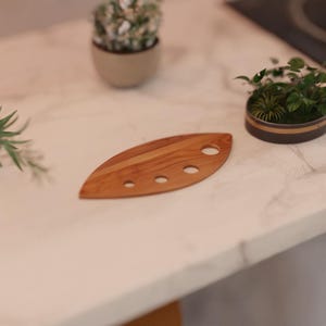 Op de afbeelding: Een houten keukengerei in de vorm van een blad met vier ronde gaten. Het keukengerei is gemaakt van lichtbruin hout en ligt op een wit marmeren aanrecht. Kleine potplanten staan op de achtergrond.
