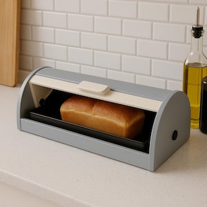 May include: A gray bread box with a cream-colored top and a black interior tray. A loaf of golden-brown bread sits inside. The box is on a white countertop, with a white tile backsplash and a bottle of oil in the background.