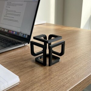 May include: A black, geometric desk organizer sits on a wooden desk next to a laptop and a notepad. The organizer has a cube-like design with open sides. Pens and pencils are in a metal holder.