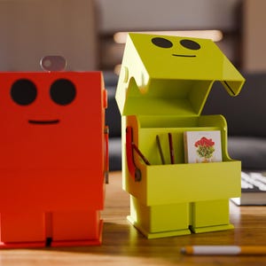 May include: Two robot-shaped storage boxes. One is bright red, the other is lime green with an open lid. Both have black eyes and a smiling mouth. The green box contains small items and a picture. A pen rests on the table.