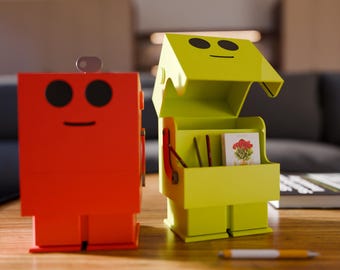 May include: Two robot-shaped storage boxes. One is bright red, the other is lime green with an open lid. Both have black eyes and a smiling mouth. The green box contains small items and a picture. A pen rests on the table.