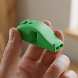 May include: A bright green whistle with a rounded top and two small holes at the end. The whistle is held in a person's hand, with a blurred background. The whistle is a simple, functional design.