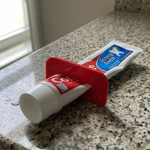 May include: A red plastic toothpaste tube squeezer with a white and red Colgate toothpaste tube. The tube is being squeezed to dispense toothpaste. The tube has the words "Strong Teeth" on it.