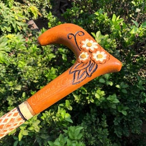 Puede incluir: Un bastón de madera con un mango curvo y un diseño floral. El mango es de color naranja cálido, con flores blancas y amarillas. El eje tiene un diseño estampado. El texto "Cheema's Co" es visible en el eje.
