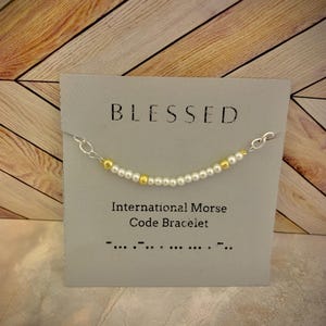 Könnte beinhalten: Eine weiße Karte mit dem Wort "BLESSED" in Schwarz gedruckt. Die Karte zeigt ein silbernes und goldenes Perlenarmband mit den Worten "International Morse Code Bracelet" und dem Morsecode für "blessed" unterhalb des Armbands.