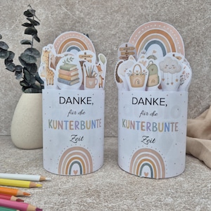 May include: Two cylindrical containers featuring the text "DANKE, für die KUNTERBUNTE Zeit" and rainbow graphics. The tops are decorated with cutouts of a giraffe, books, pencils, a basket, a backpack, and a cloud with hanging stars.