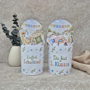 Könnte beinhalten: Zwei zylindrische weiße Behälter mit Blumenmustern und schulbezogenen Illustrationen wie einer Tafel, einer Farbpalette und Büchern. Der linke Behälter trägt die Aufschrift Endlich Schulkind, der rechte die Aufschrift Du bist 1. Klasse.