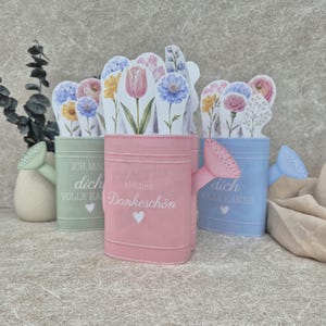 Peut inclure: Trois arrosoirs de couleur pastel en vert clair, rose et bleu clair, chacun rempli de découpes en papier représentant diverses fleurs. L'arrosoir rose porte le texte "kleines Dankeschön" et une petite icône de cœur blanc.