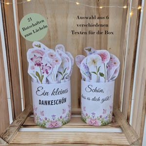 Puede incluir: Dos recipientes cilíndricos blancos con diseños florales y texto. Cada uno contiene tarjetas con forma de flor con ilustraciones florales. Uno dice "Ein kleines DANKESCHÖN" y el otro dice "Schön, dass es dich gibt!".
