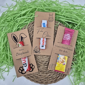 Könnte beinhalten: Drei Oster-Geschenkkarten mit Schokoladen-Leckereien. Die Karten sind braun mit Hasen- und Eier-Designs und Text auf Deutsch. Die Schokoladen sind in bunten Verpackungen. Die Karten sind auf einer gewebten Matte mit grünem Papiergras angeordnet.