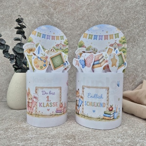 May include: Two cylindrical white containers featuring school-themed illustrations. The left container shows a rabbit and the text 'Du bist 1. KLASSE'. The right container shows a fox and the text 'Endlich SCHULKIND'. Both feature colorful school supplies.