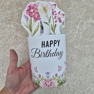 May include: A cylindrical Happy Birthday greeting card featuring a white base with black text, decorated with pink and purple floral illustrations, green foliage, and small butterflies. The top edge displays several die-cut paper flowers.