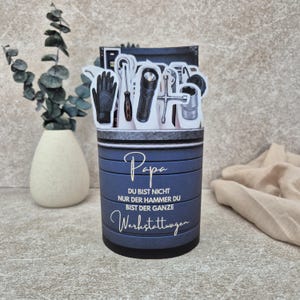 May include: A cylindrical blue container featuring a graphic of various tools including a glove, screwdriver, flashlight, and wrench. The text reads: Papa DU BIST NICHT NUR DER HAMMER DU BIST DER GANZE Werkstattwagen. The container has a dark base.
