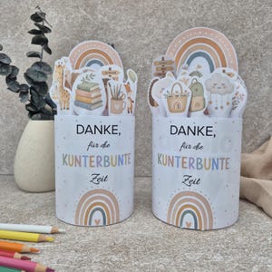 Può includere: Due contenitori cilindrici con il testo "DANKE, für die KUNTERBUNTE Zeit" e grafiche arcobaleno. Le parti superiori sono decorate con ritagli di una giraffa, libri, matite, un cestino, uno zaino e una nuvola con stelle pendenti.