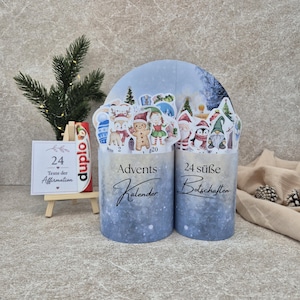 May include: A two-part cylindrical advent calendar with a blue and white snowy gradient finish. The front features the text Advents Kalender and 24 süße Botschaften, topped with various Christmas-themed paper cutouts including a gingerbread man.